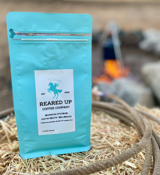 Turquoise bag of Reared of Coffee Cowboy Blend on a hay bale, surrounded by a lasso, in front of an open fire with a cowboy coffee pot over the fire.