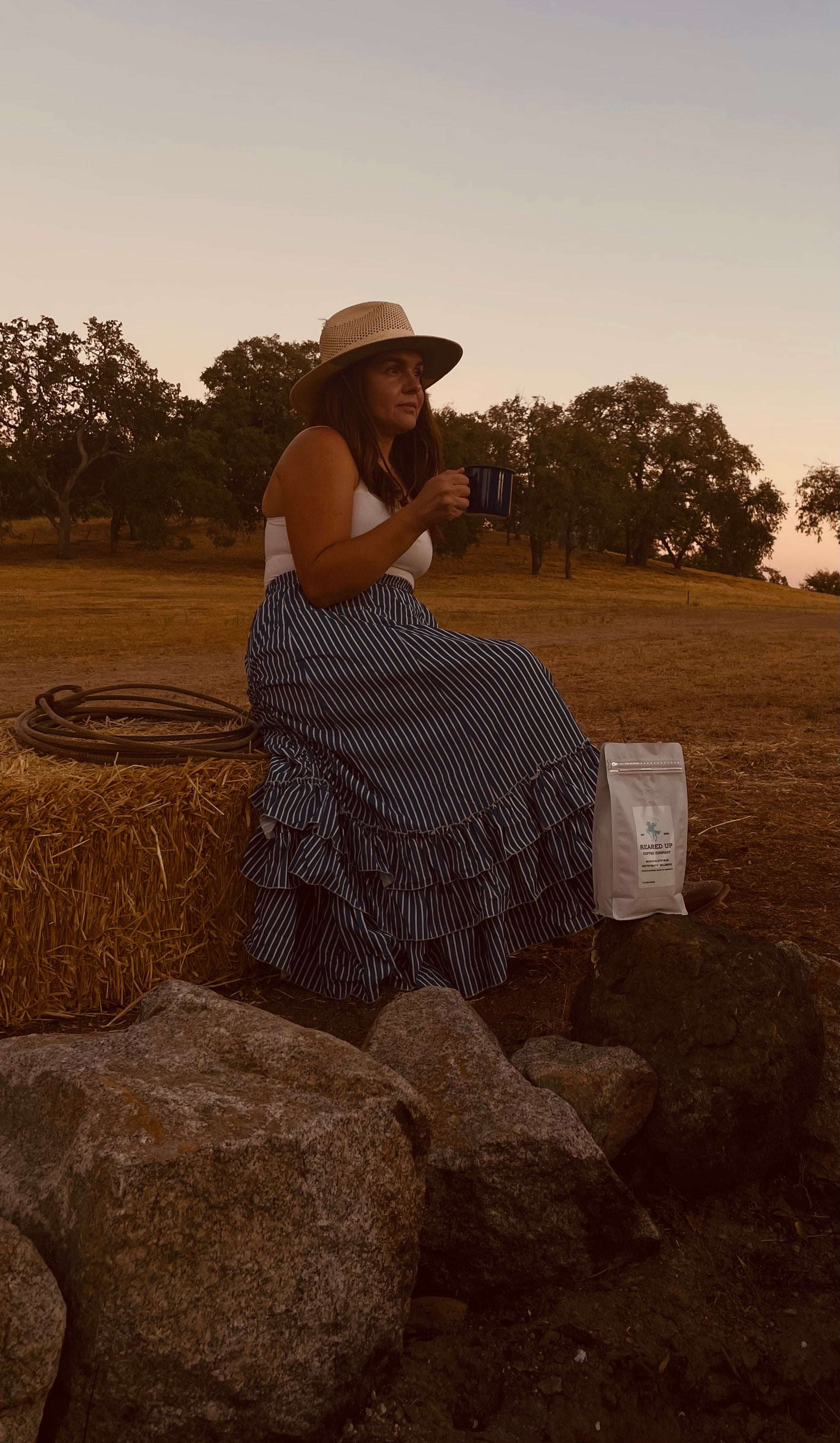 Jennifer Garcia sitting on a bale of hay, drinking coffee, with a white bag of Reared Up Coffee Company.